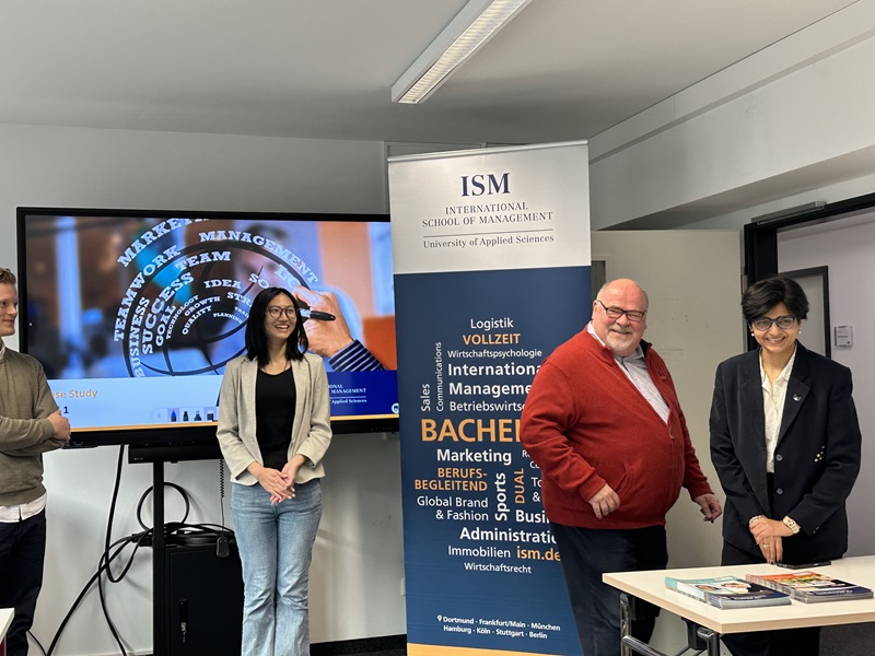 In einem Seminarraum der Euro Akademie Wiesbaden stehen drei Personen vor einer Präsentationsfläche und einem Roll-up-Banner der International School of Management (ISM). Links steht eine junge Frau in heller Jacke und Jeans lächelnd vor einem großen Bildschirm, auf dem eine Grafik zu Themen wie Management, Teamwork und Erfolg zu sehen ist. Rechts daneben stehen zwei weitere Personen, ein Mann in rotem Pullover und eine Frau im dunklen Blazer, beide freundlich zugewandt. Auf dem Tisch vor ihnen liegen verschiedene Bücher und Materialien. Die Szene entstand während eines Workshops der ISM zum Thema Management Erleben für Studierende und vermittelt eine lebendige, praxisorientierte Lernatmosphäre.