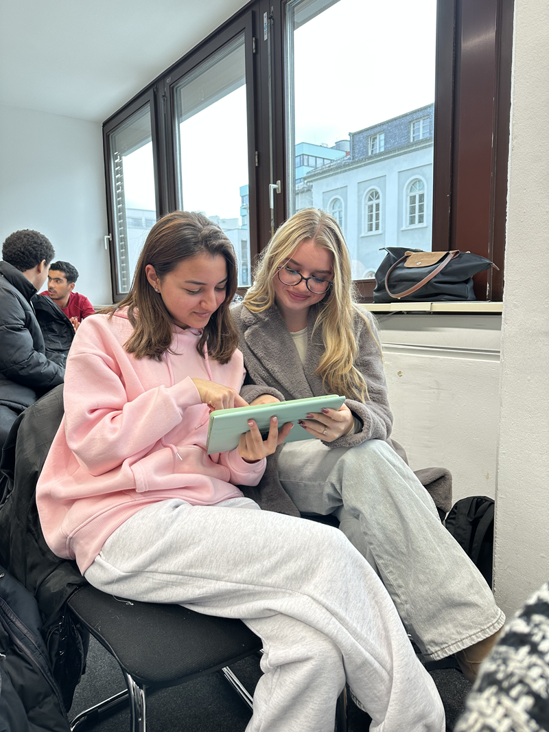 Zwei junge Frauen sitzen nebeneinander auf schwarzen Stühlen in einem hellen Raum mit großen Fenstern, durch die ein historisches Gebäude zu sehen ist. Die Frau links trägt einen hellrosa Pullover und eine hellgraue Hose, die Frau rechts einen grauen Pullover und eine hellgraue Hose. Beide konzentrieren sich auf ein hellgrünes Tablet, das die linke Frau hält, während die rechte Frau mit der rechten Hand darauf zeigt. Im Hintergrund sind weitere Personen zu erkennen, die ebenfalls auf Stühlen sitzen. Auf der Fensterbank liegt eine schwarze Tasche mit braunen Griffen. Die Szene vermittelt eine Lern- oder Workshop-Atmosphäre, passend zum Thema "Management Erleben" im Rahmen eines Workshops der ISM an der Euro Akademie Wiesbaden.
