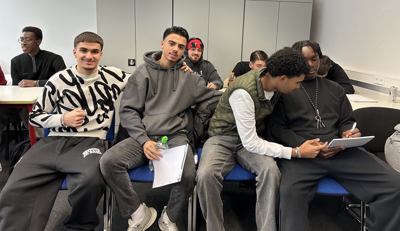 In einem Seminarraum der Euro Akademie Wiesbaden sitzen mehrere Studierende auf blauen Stühlen nebeneinander. Sie tragen legere Kleidung, darunter Pullover, Hoodies und Jacken in gedeckten Farben. Einige halten Notizblätter oder ein Tablet in den Händen. Die Studierenden sind in kleinen Gruppen zusammengesetzt, wobei zwei von ihnen sich eng nebeneinandersitzend über das Tablet austauschen. Im Hintergrund sind weiße Schränke an der Wand zu sehen. Die Szene vermittelt eine konzentrierte und interaktive Lernatmosphäre im Rahmen eines Workshops der ISM zum Thema "Management Erleben".