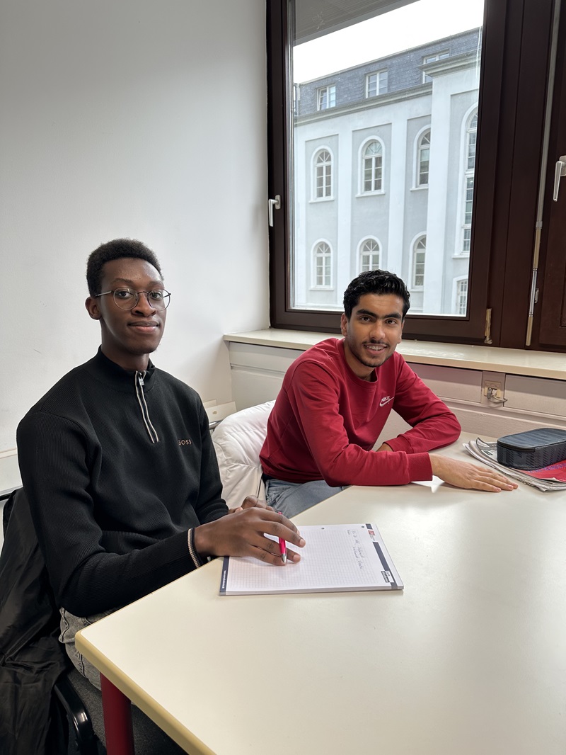 Zwei Studierende sitzen an einem hellen Tisch in einem hellen Raum mit großen Fenstern, durch die ein historisches Gebäude mit weißen Fassaden und hohen Fenstern sichtbar ist. Der Studierende links trägt einen schwarzen Pullover mit Reißverschluss und hält einen roten Stift über einem Notizblock, der auf dem Tisch liegt. Der Studierende rechts trägt ein rotes Sweatshirt und hat die Arme auf dem Tisch abgestützt. Auf dem Tisch liegen außerdem ein schwarzes Etui und ein rotes Buch. Die Szene zeigt eine ruhige Lernatmosphäre während eines Workshops zum Thema "Management Erleben" an der Euro Akademie Wiesbaden im Rahmen der ISM-Studienveranstaltung.