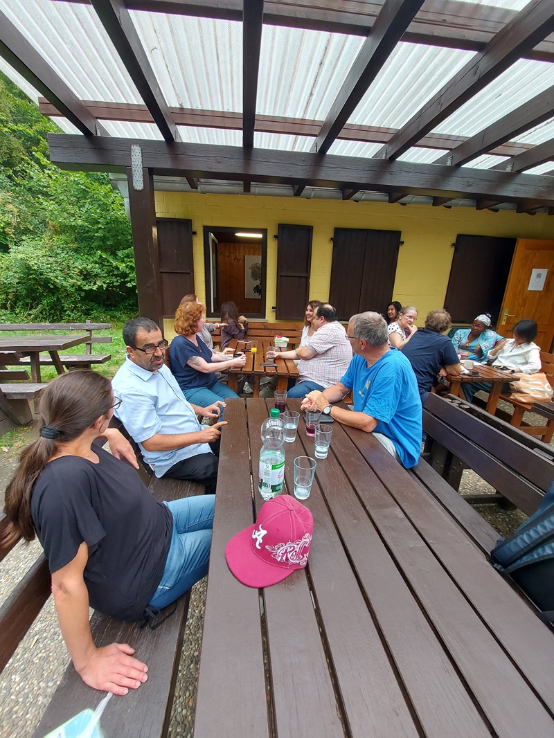 Mehrere Mitarbeitende sitzen an Holztischen unter einer überdachten Terrasse und unterhalten sich. Auf den Tischen stehen Gläser, Flaschen und Tassen. Im Hintergrund befindet sich ein gelb gestrichenes Gebäude mit dunklen Fensterläden, umgeben von dichtem Grün.