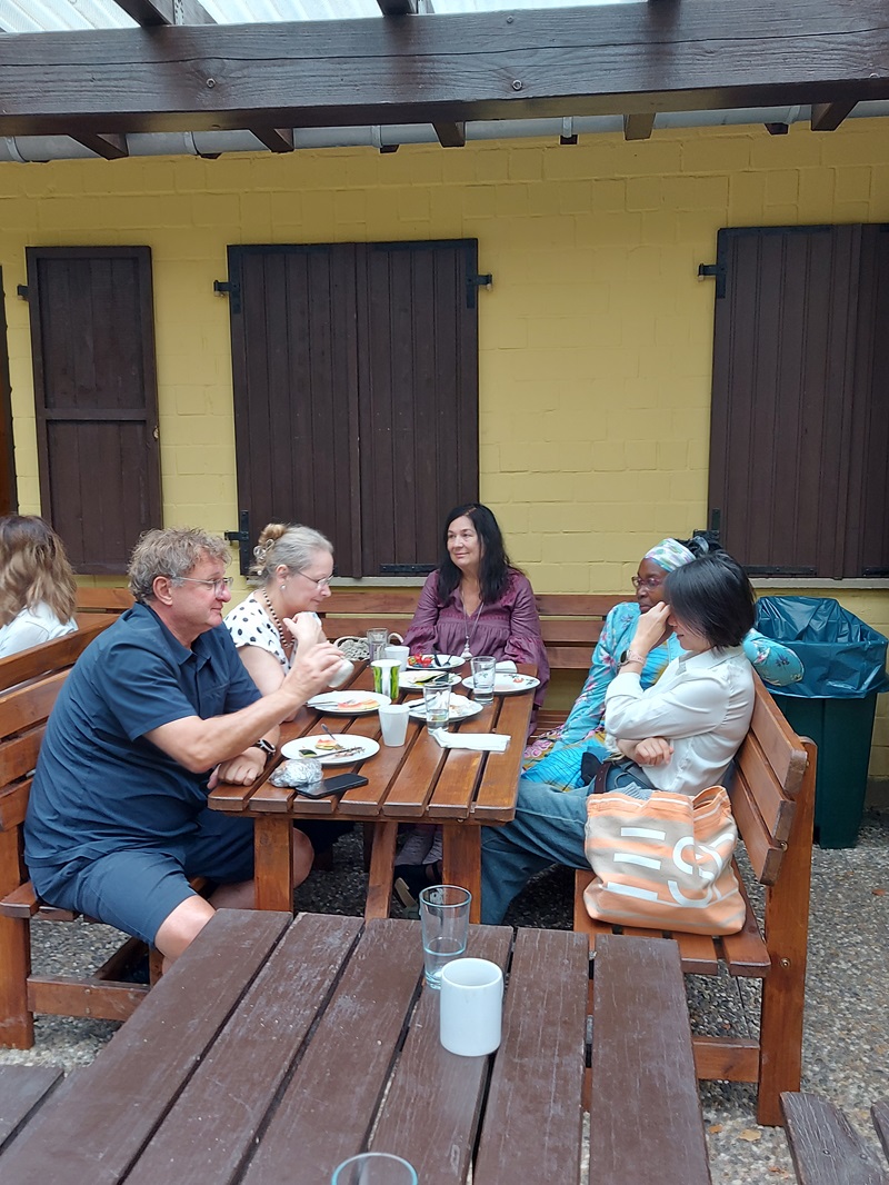 Fünf Mitarbeitende sitzen an einem Holztisch auf einer überdachten Terrasse und unterhalten sich während einer gemeinsamen Mahlzeit. Auf dem Tisch stehen Teller, Gläser und Tassen. Im Hintergrund ist eine gelbe Gebäudewand mit dunklen Fensterläden zu sehen.