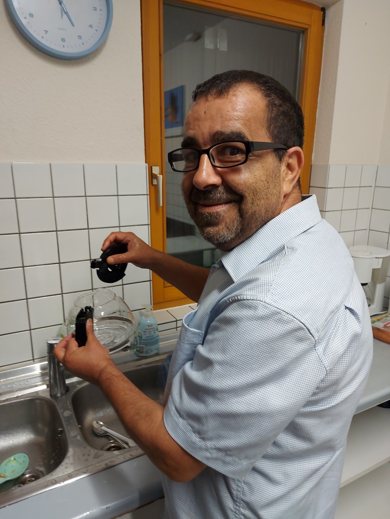 Ein Mitarbeiter mit Brille und kurzem Bart steht lächelnd an einem Spülbecken in einer Küche. Er trägt ein helles Hemd und hält eine Glaskanne sowie einen schwarzen Deckel in den Händen. Im Hintergrund sind weiße Fliesen, ein Fenster mit gelbem Rahmen und eine Wanduhr zu sehen. Der Mann blickt lächelnd über die Schulter zur Kamera.