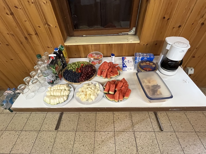 Auf einem rechteckigen Tisch stehen verschiedene Lebensmittel und Getränke. In der Mitte befinden sich mehrere Teller mit geschnittenem Obst, darunter Wassermelone, Trauben, Birnen und Bananen. Links stehen mehrere Flaschen, darunter Wasser und Saft, sowie Gläser. Rechts sind eine Kaffeemaschine, eine Packung Milch, eine Schale mit Keksen und ein Behälter mit Gebäck zu sehen. Der Tisch steht vor einer Holzvertäfelung und einem Fenster.