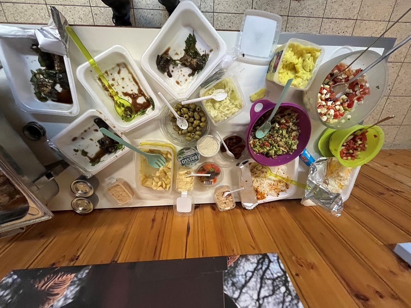 Verschiedene Schalen und Behälter mit Salaten, Oliven, Käse, Saucen und weiteren Lebensmitteln stehen auf einem Tisch. Einige Schalen sind aus Kunststoff, andere aus Metall. Besteck wie Löffel und Zangen liegen in den Behältern. Die Oberfläche des Tisches ist teilweise aus Holz, daneben sind Fliesen zu sehen. Die Szene zeigt eine Auswahl an Speisen, die zum Selbstbedienen bereitgestellt sind.