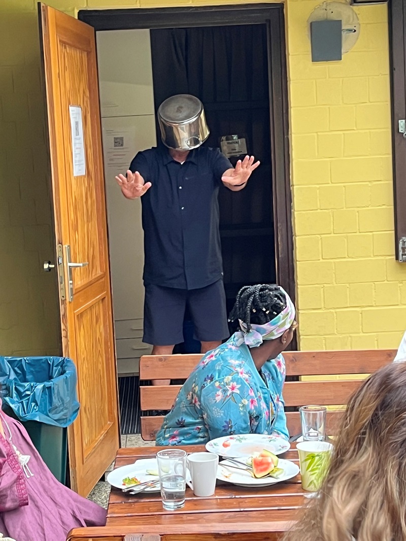 Ein Mitarbeiter steht in einem Türrahmen und trägt einen Kochtopf verkehrt herum auf dem Kopf. Die Arme sind nach vorne ausgestreckt. Im Vordergrund sitzen mehrere Menschen an einem Holztisch, auf dem Teller mit Essensresten, Gläser und Besteck stehen. Die Szene spielt sich vor einer gelben Wand ab.