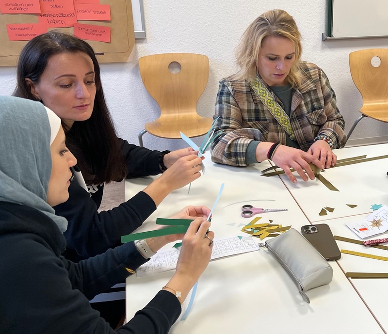 Drei Schülerinnen, darunter eine mit Kopftuch, basteln gerade an ihren Fröbelsternen. Auf dem Tisch liegen Anleitungen, eine Schere, und ein Handy sowie einige Papierschnipsel.