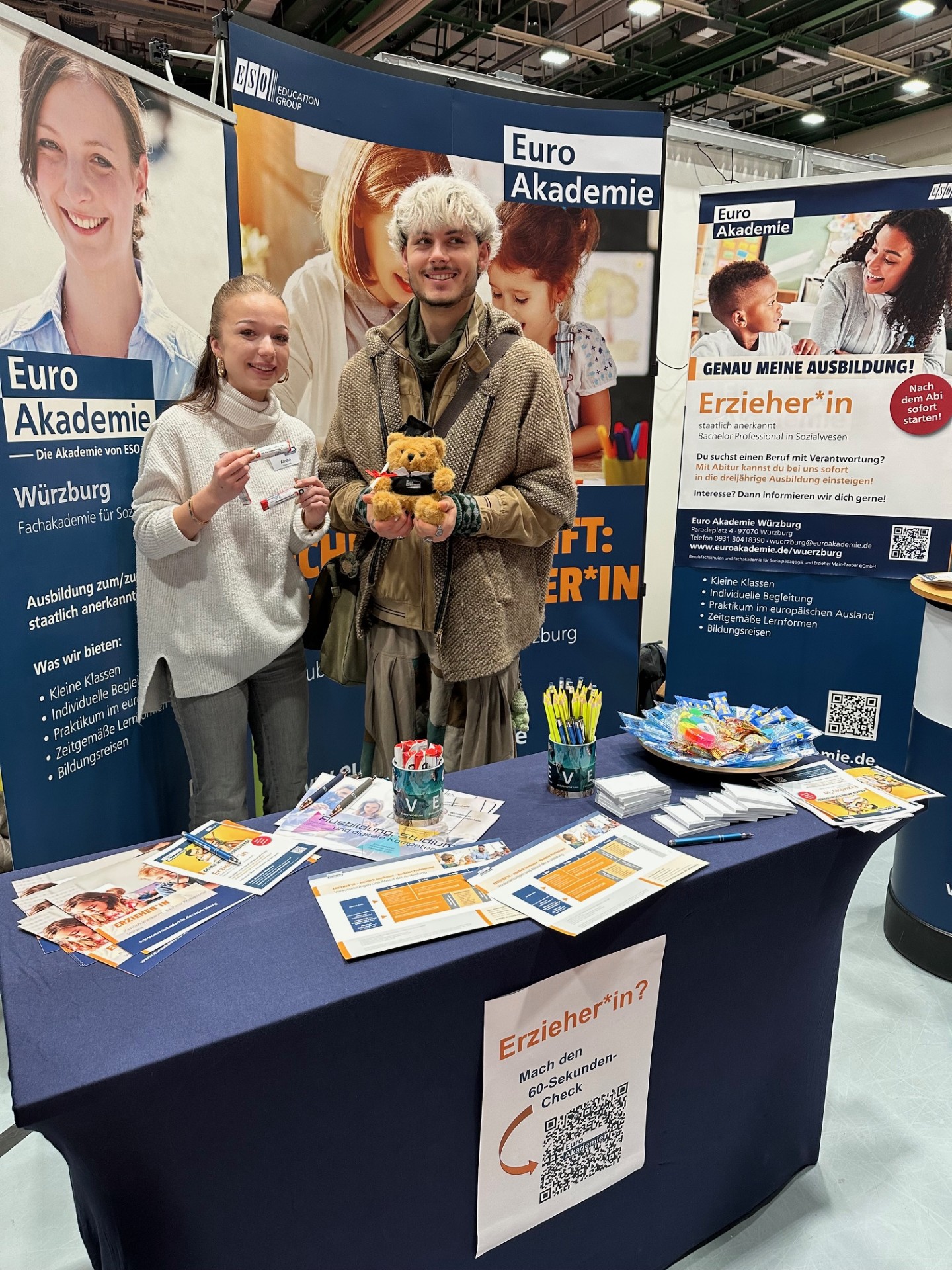 Zwei Personen stehen an einem Messestand der Euro Akademie vor großen blauen und weißen Infowänden, halten einen kleinen Teddybären mit Doktorhut und einen Giveaway-Stift in der Hand.