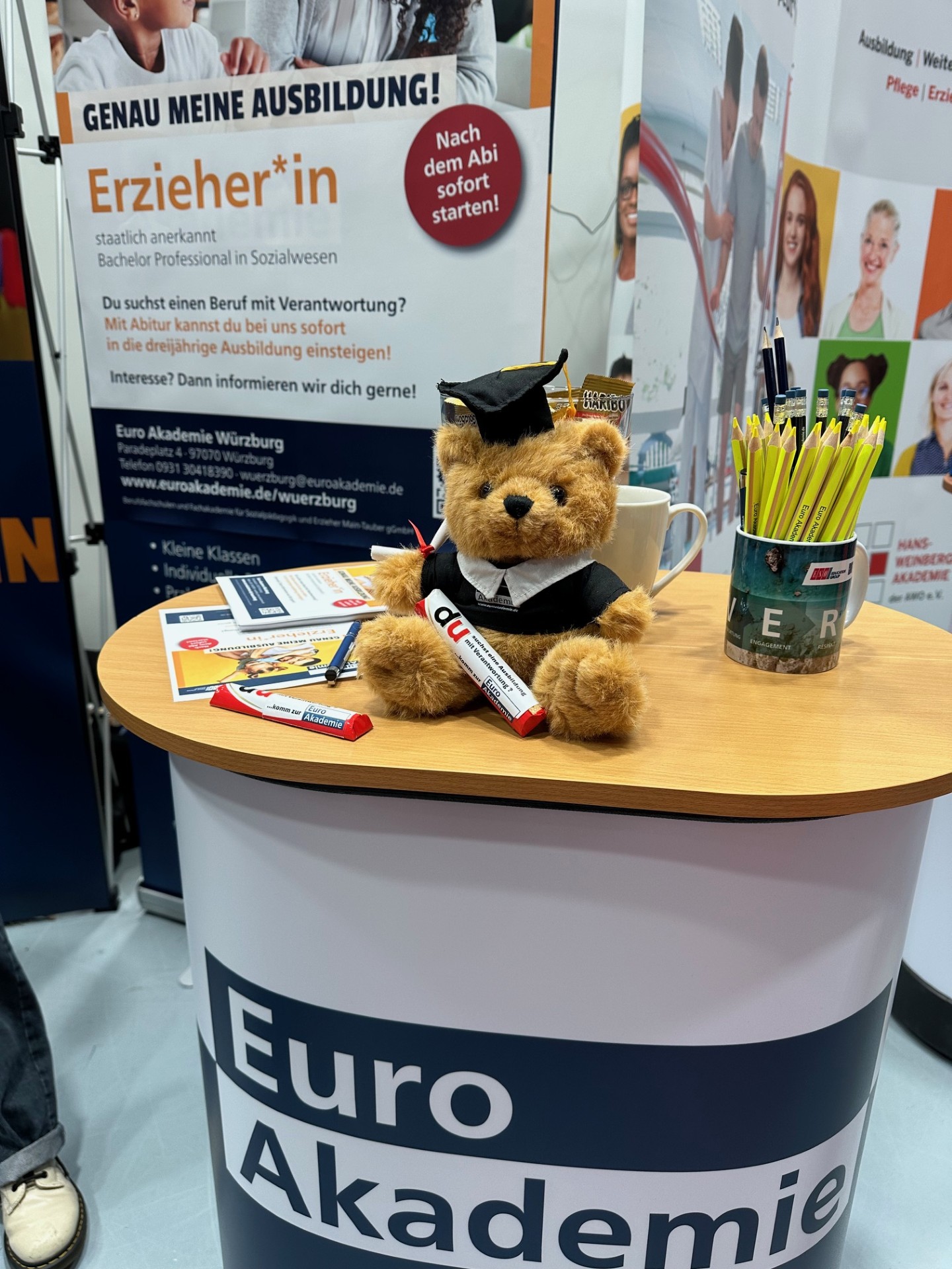 Auf einem Messestand der Euro Akademie sitzt ein kleiner Teddybär mit Doktorhut und Robe auf einem runden Tresen, umgeben von Flyern, Stiften und einer Tasse mit Bleistiften; im Hintergrund werben Roll‑up‑Banner auf Deutsch für die Ausbildung zur Erzieherin bzw. zum Erzieher.