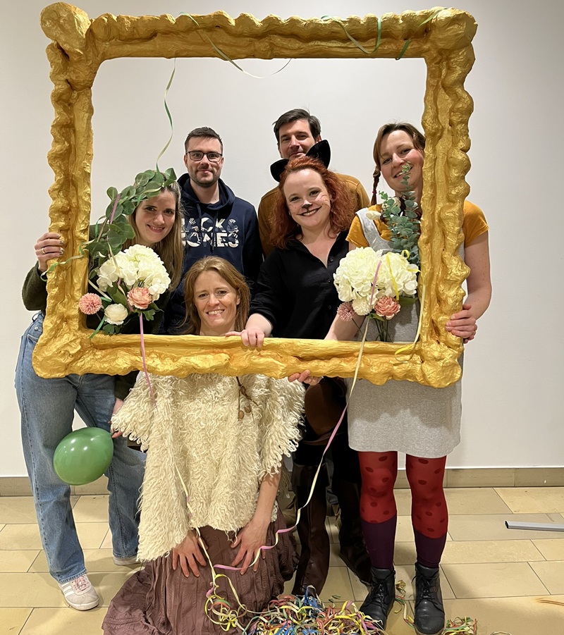 Lehrkraft und Studierende schauen lächelnd in ihren Faschingskostümen durch einen übergroßen selbst gestalteten Bilderrahmen und posieren für ein Foto.