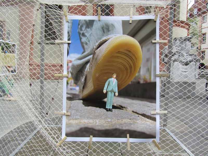 Blick auf ein an der Installation am Netz mit Holzklammern angebrachtes Bild in Close Up, auf dem eine Miniaturfigut eines Mannes im Anzug abgebildet ist, der andeutungsweise von einem riesigen Schuh platt getreten werden wird. 