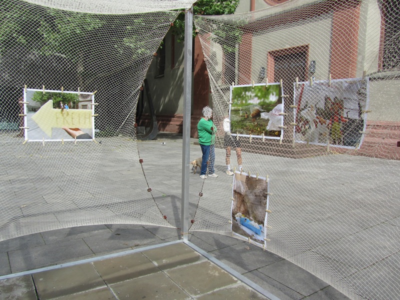 Blick auf die Installation in Würfelform, die an vier Seiten mit einem Netz bespannt ist, an dem Bilder mit Holzklammern angebracht sind. Im Hintergrund stehen zwei Menschen, mit kleinem Hund. .