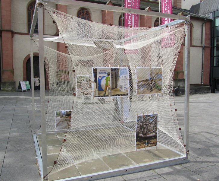 Blick auf die begehbare Installation in Würfelform, an der unterschiedliche Bilder hängen, unter anderem eines, dass einen Jungen mit Milchkanne zeigt, der an einer Werbetafel für haltbare Vollmilch vorbeigeht. 