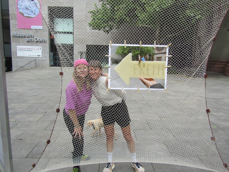 Blick auf die Installation und das gespannte Netz, an dem eini Bild angebracht ist, auf dem zwei kleine Figuren auf einem großen gelben Pfeil sitzen, auf dem Freiheit steht. Hinter dem Netz sind zwei lächelnde Frauen zu sehen, die in farbenfrohen Outfits in die Kamera lächeln. 