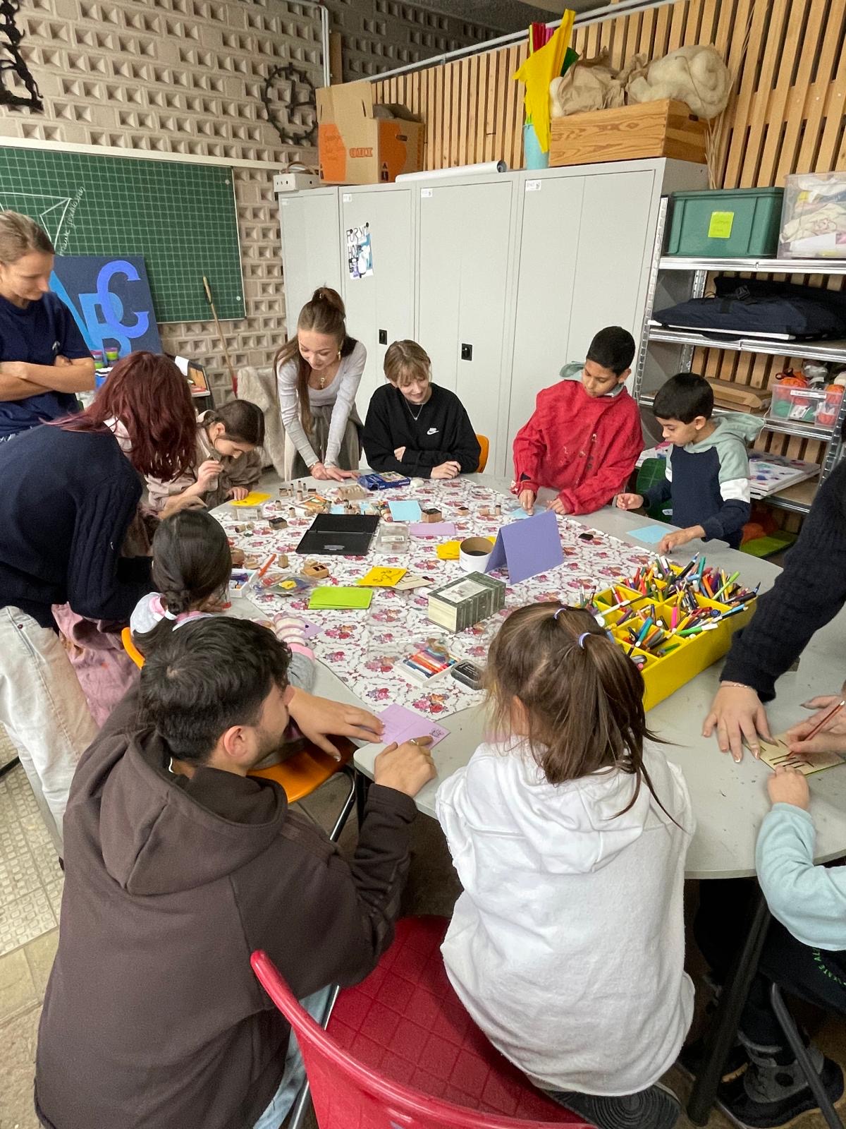 Gruppe von Kindern und Anleitenden arbeitet an einem großen Bastel- und Drucktisch mit Stempeln, Papieren und Stiften im Klassenraum.