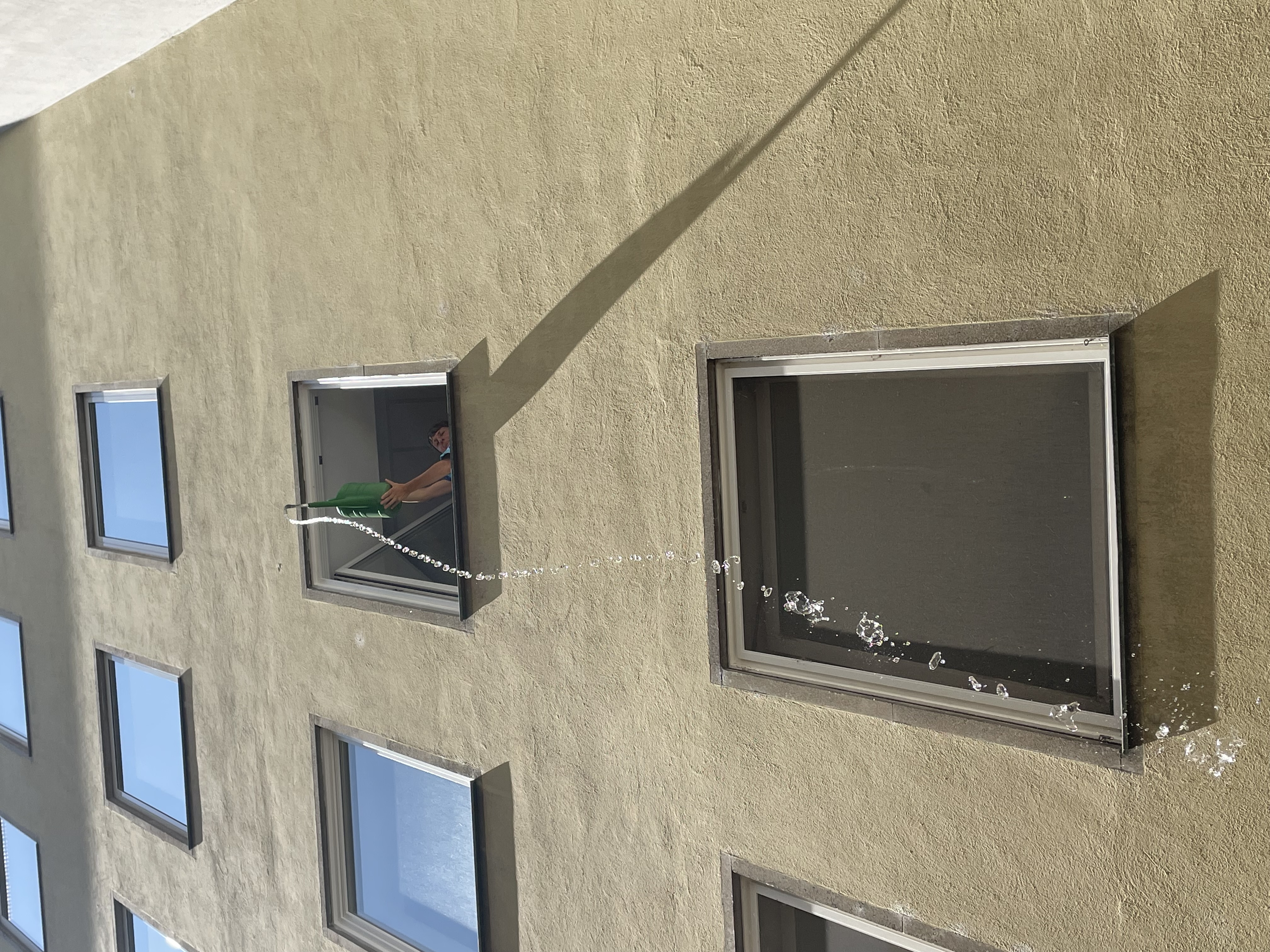 Wasser-Quiz und Wasserschlacht Eine Person gießt Wasser mit einer grünen Gießkanne aus einem Fenster in einem oberen Stockwerk eines mehrstöckigen Hauses. Der Wasserstrahl fällt in einem Bogen nach unten.
