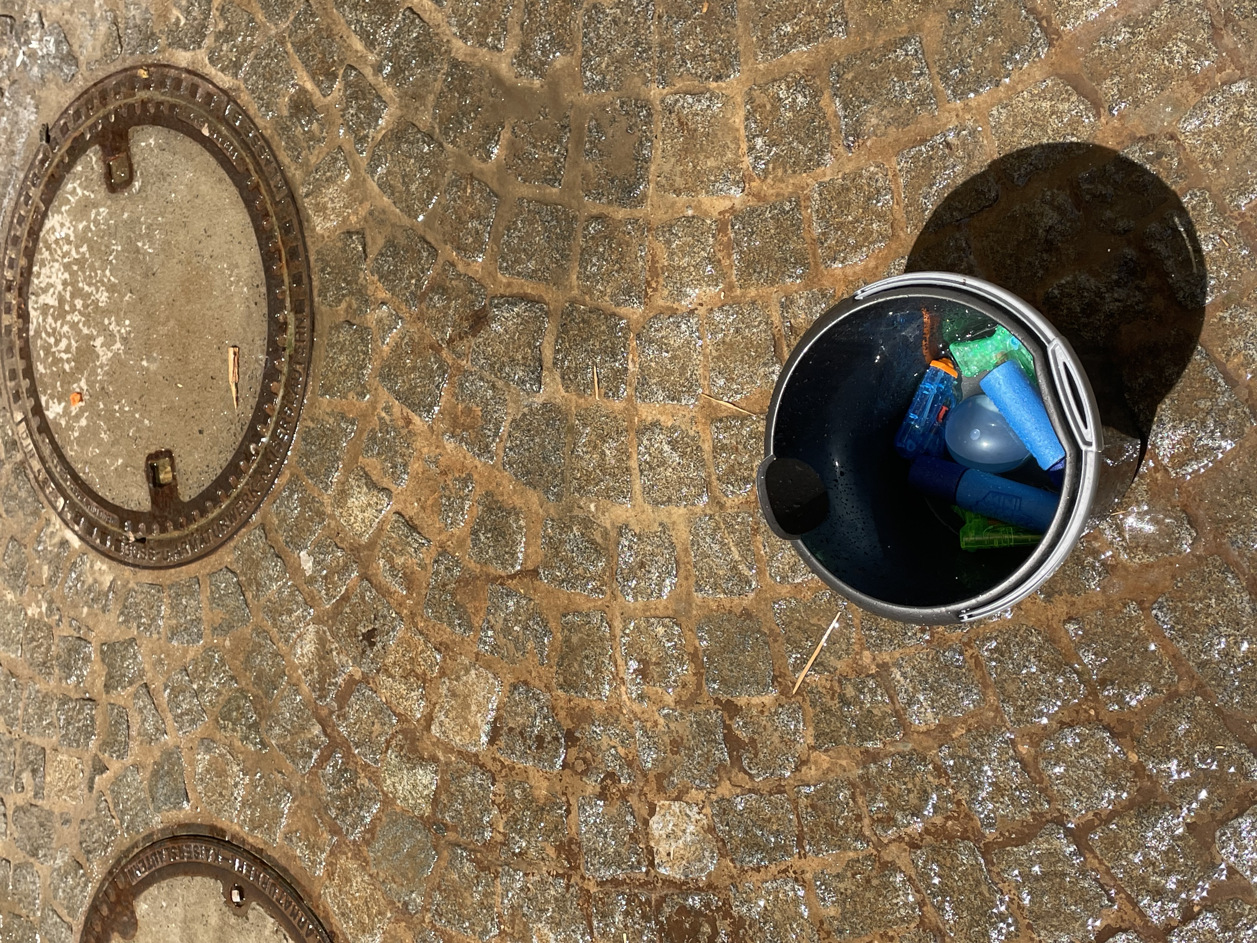 Wasser-Quiz und Wasserschlacht Ein schwarzer Eimer steht auf einem nassen Kopfsteinpflaster. Darin befinden sich bunte Wasserpistolen und Wasserbomben. Im Hintergrund sind zwei Kanaldeckel zu sehen.