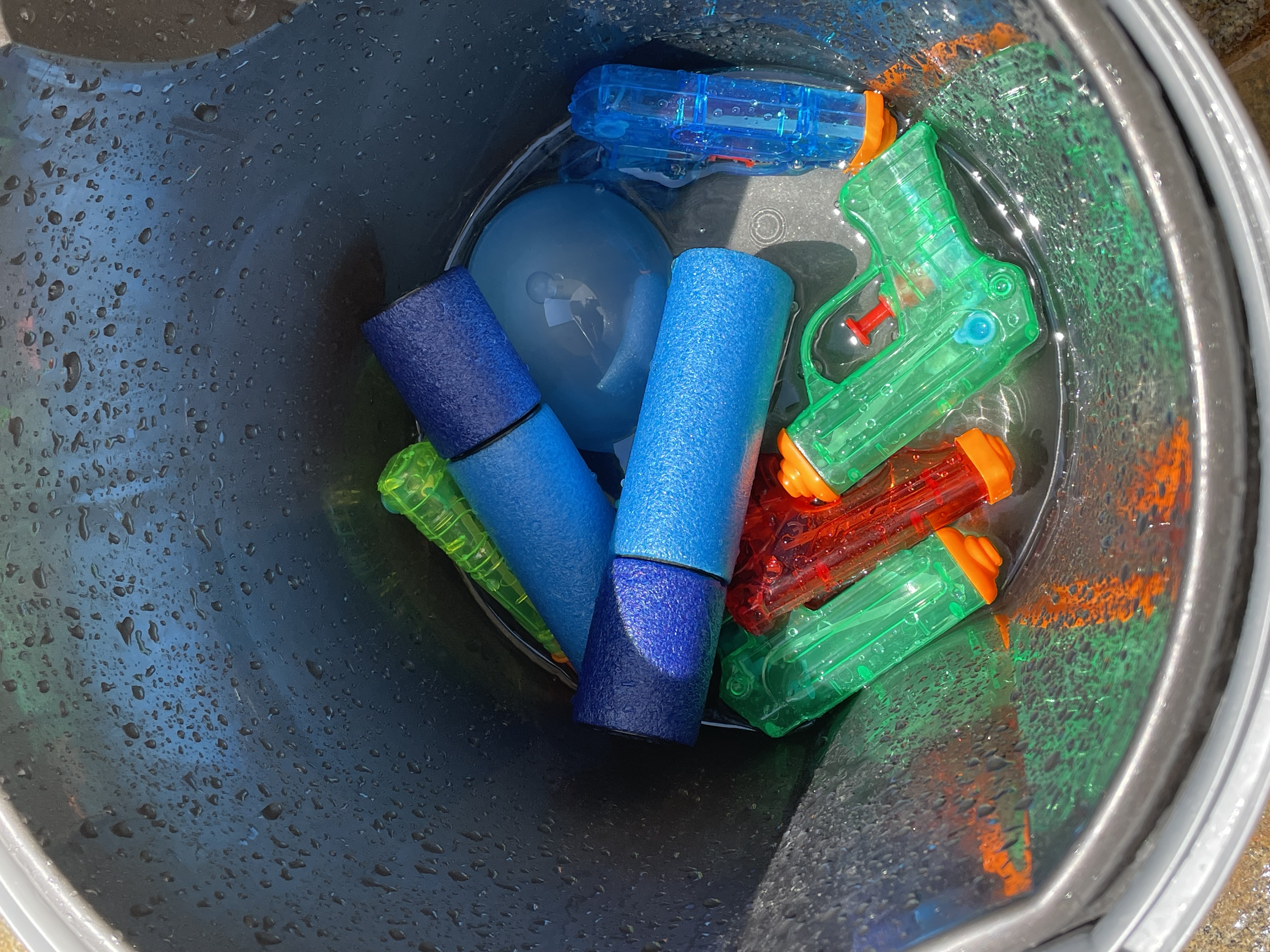 Wasser-Quiz und Wasserschlacht Nahaufnahme des schwarzen Eimers mit mehreren bunten Wasserpistolen, zwei blauen Schaumstoff-Wasserspritzen und einer gefüllten Wasserbombe.