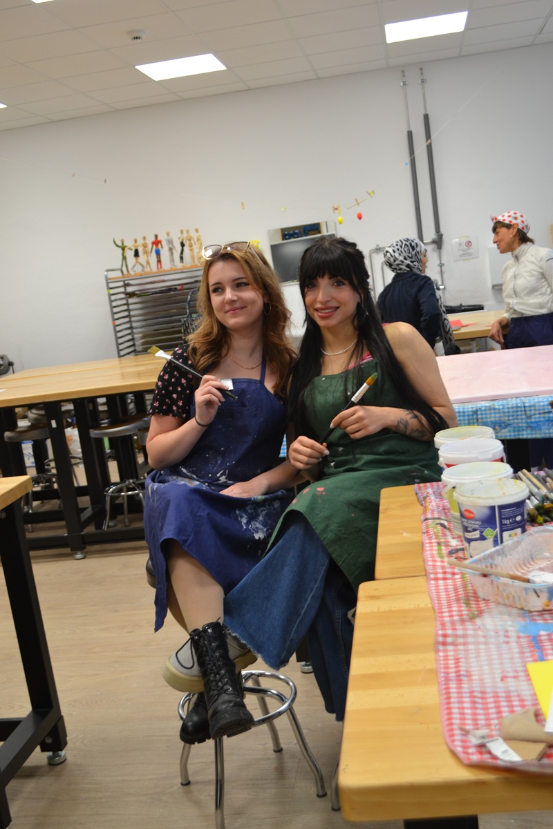Zwei Teilnehmerinnen des Tages der offenen Tür an der Euro Akademie Würzburg posieren lächelnd, auf Stühlen im Atelier sitzend, mit Malschürzen und Pinsel in der Hand für ein Bild. 