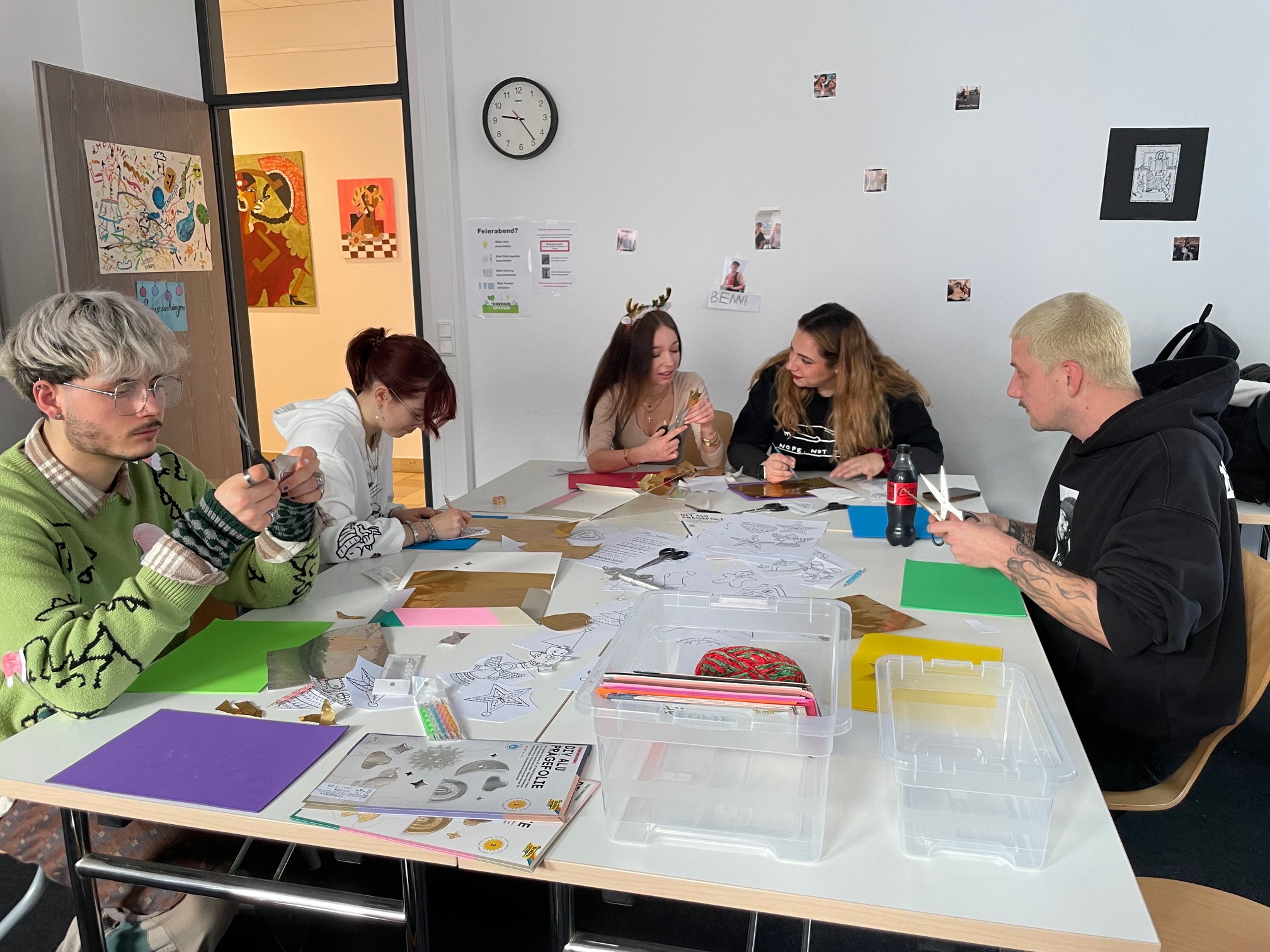 Mehrere Studierende arbeiten konzentriert an einem großen Tisch mit farbigem Papier, Vorlagen und Bastelmaterialien, im Hintergrund hängen kleine Fotos an der Wand.