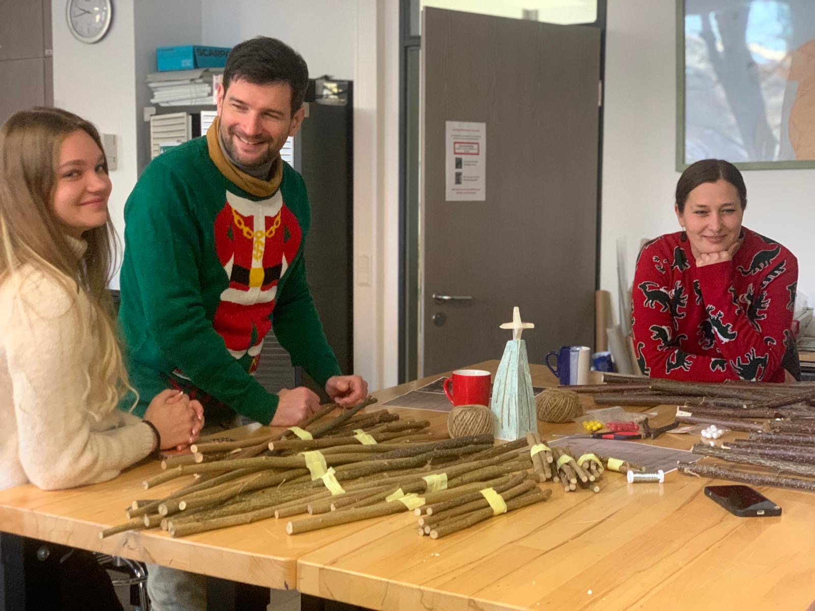 Drei Personen in weihnachtlichen Pullis stehen an einer Werkbank und lachen, vor ihnen liegen viele Holzstäbe und Bastelmaterial für dekorative Weihnachtsbäume.