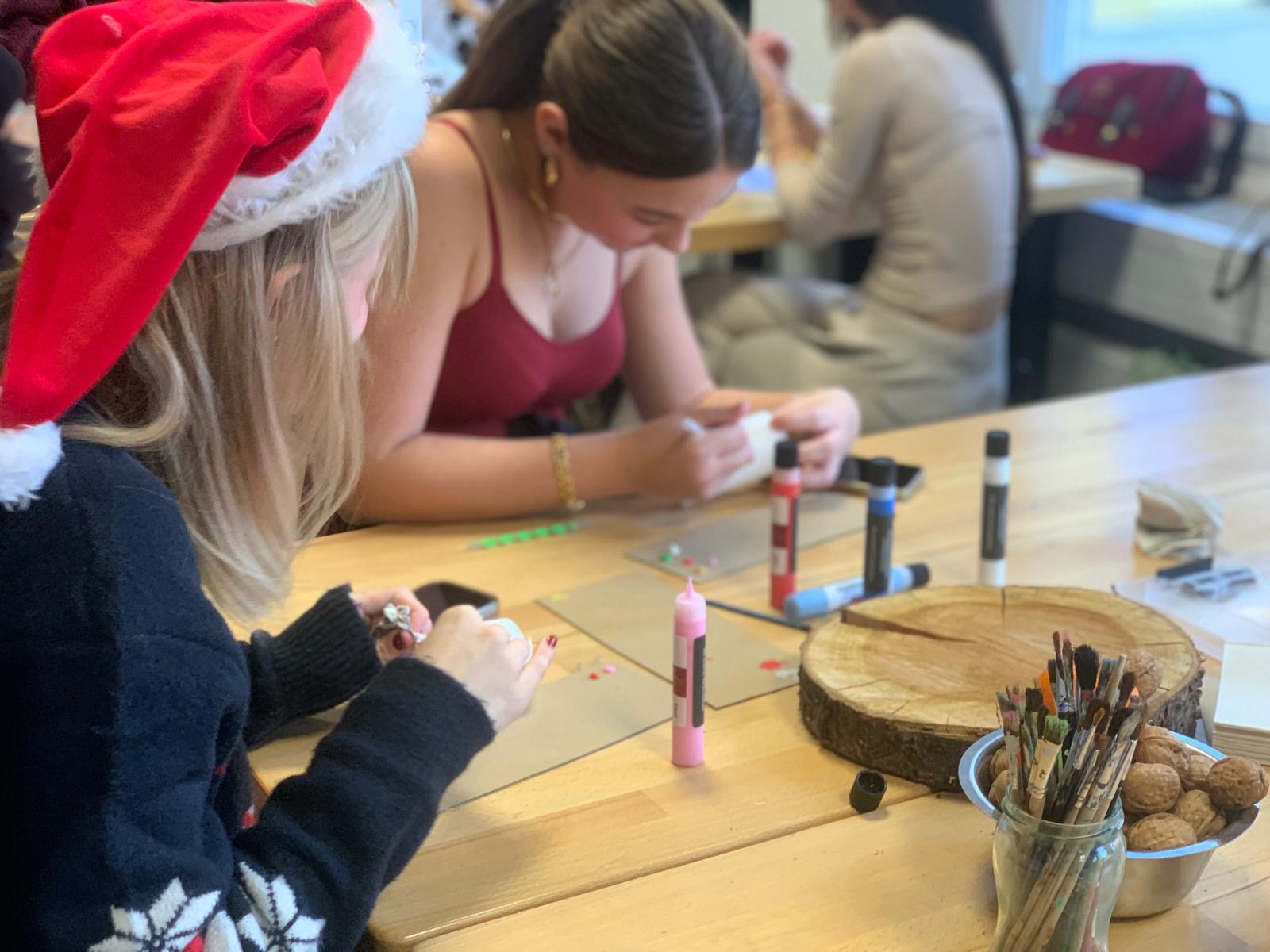 Zwei junge Frauen sitzen an einem Tisch mit Pinseln, Farben und Holzscheiben; eine trägt eine Nikolausmütze, beide verzieren konzentriert kleine Weihnachtsprojekte.