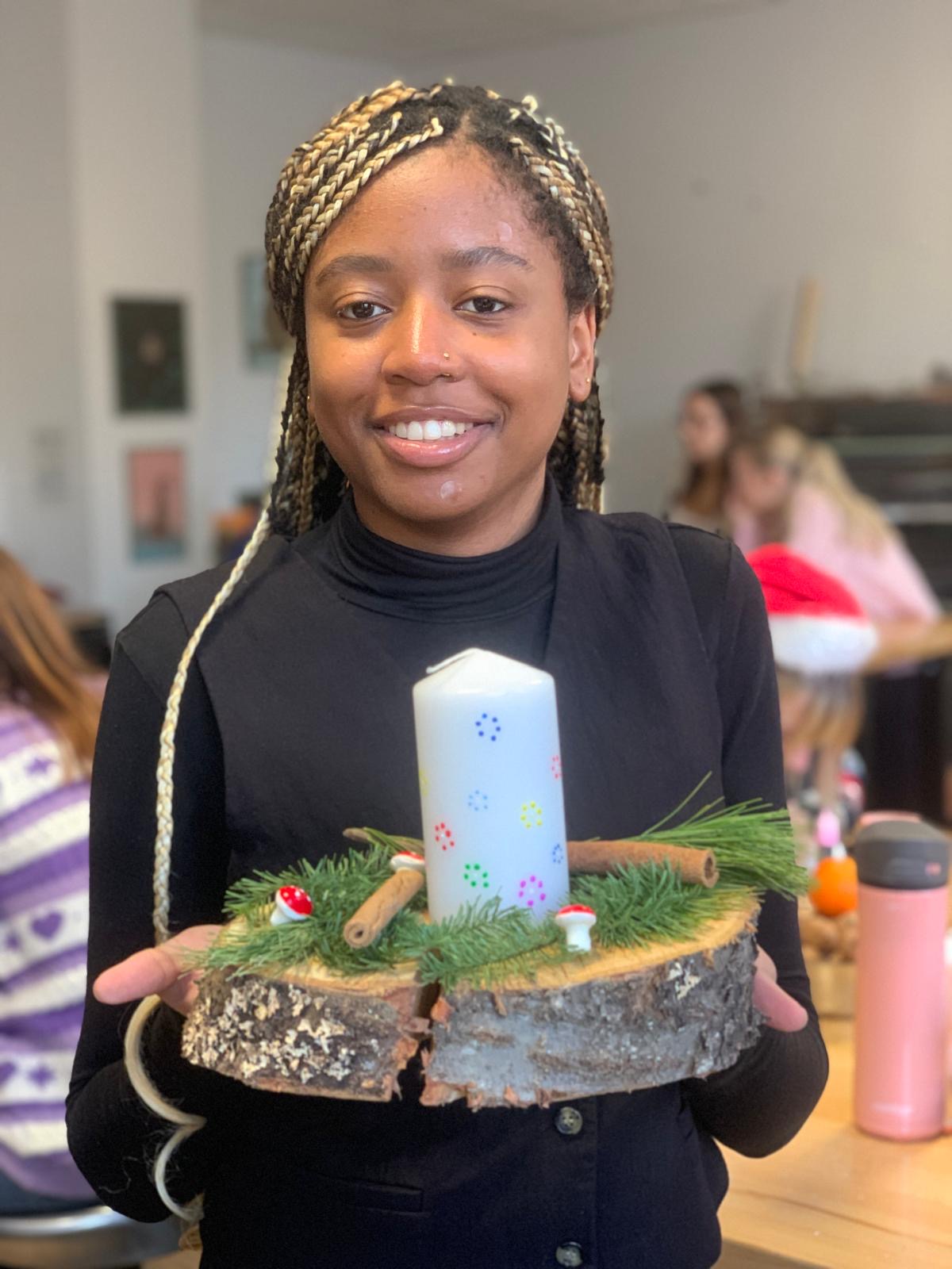 Eine Schülerin hält lächelnd ein selbst gestaltetes Kerzengesteck aus einer Holzscheibe mit Tannengrün, Zimtstangen und einer dekorierten weißen Kerze in die Kamera.