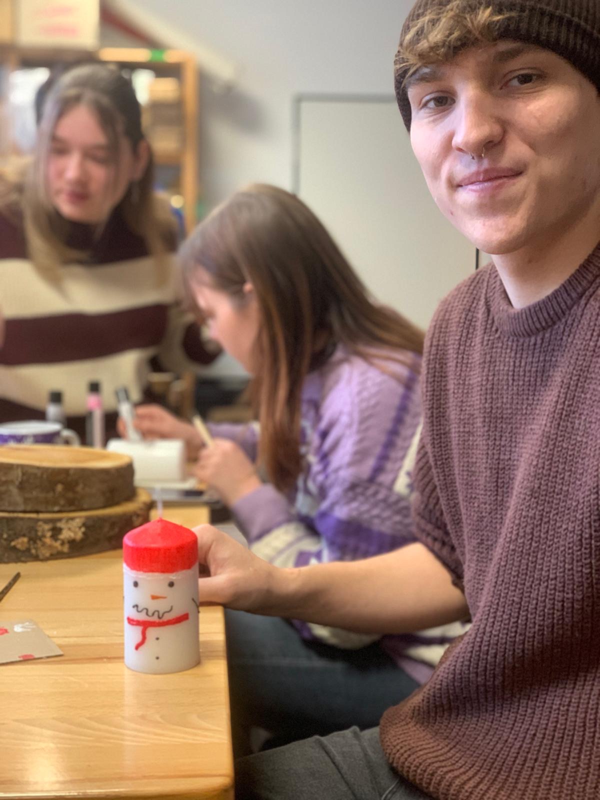 Ein Student zeigt stolz eine von ihm bemalte Kerze im Schneemann-Design, während hinter ihm weitere Teilnehmende an ihren Weihnachtskerzen arbeiten.
