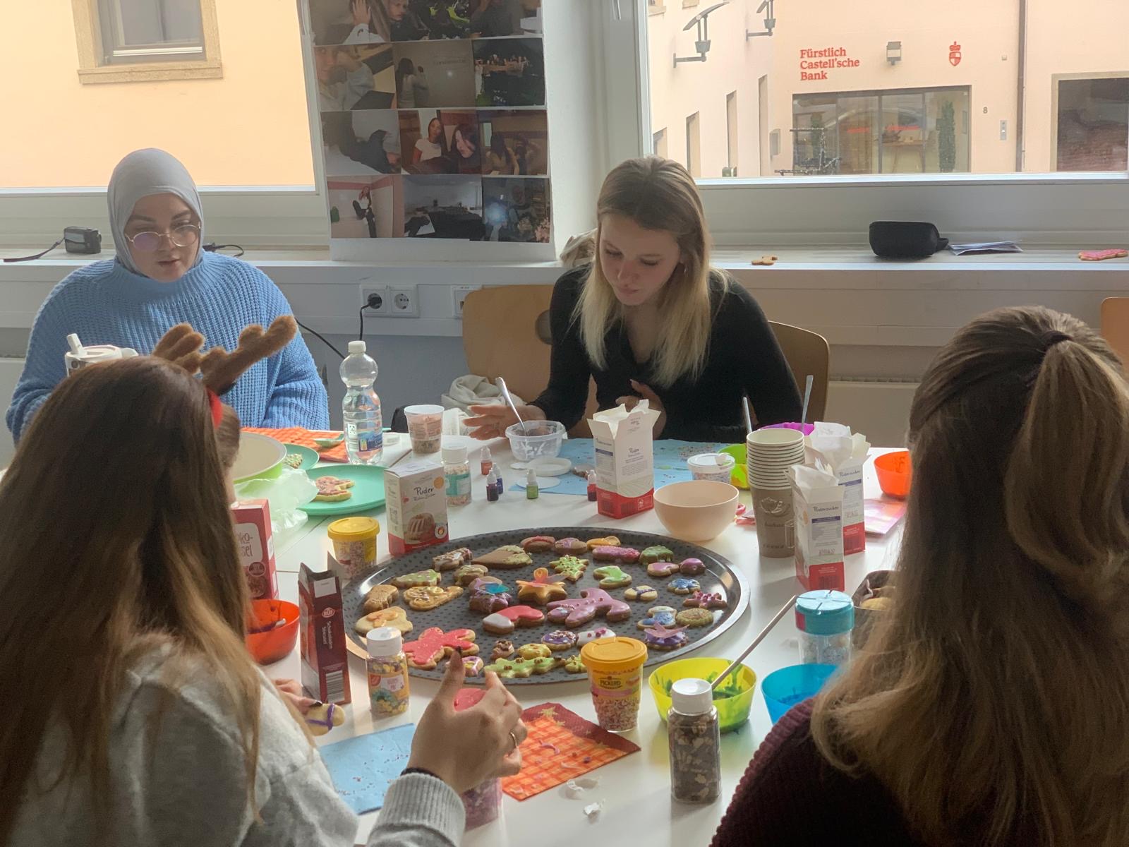 Mehrere Studierende sitzen um einen Tisch und verzieren gemeinsam Weihnachtsplätzchen mit Zuckerguss und Streuseln, in der Mitte steht ein großes Blech voller bunter Kekse.