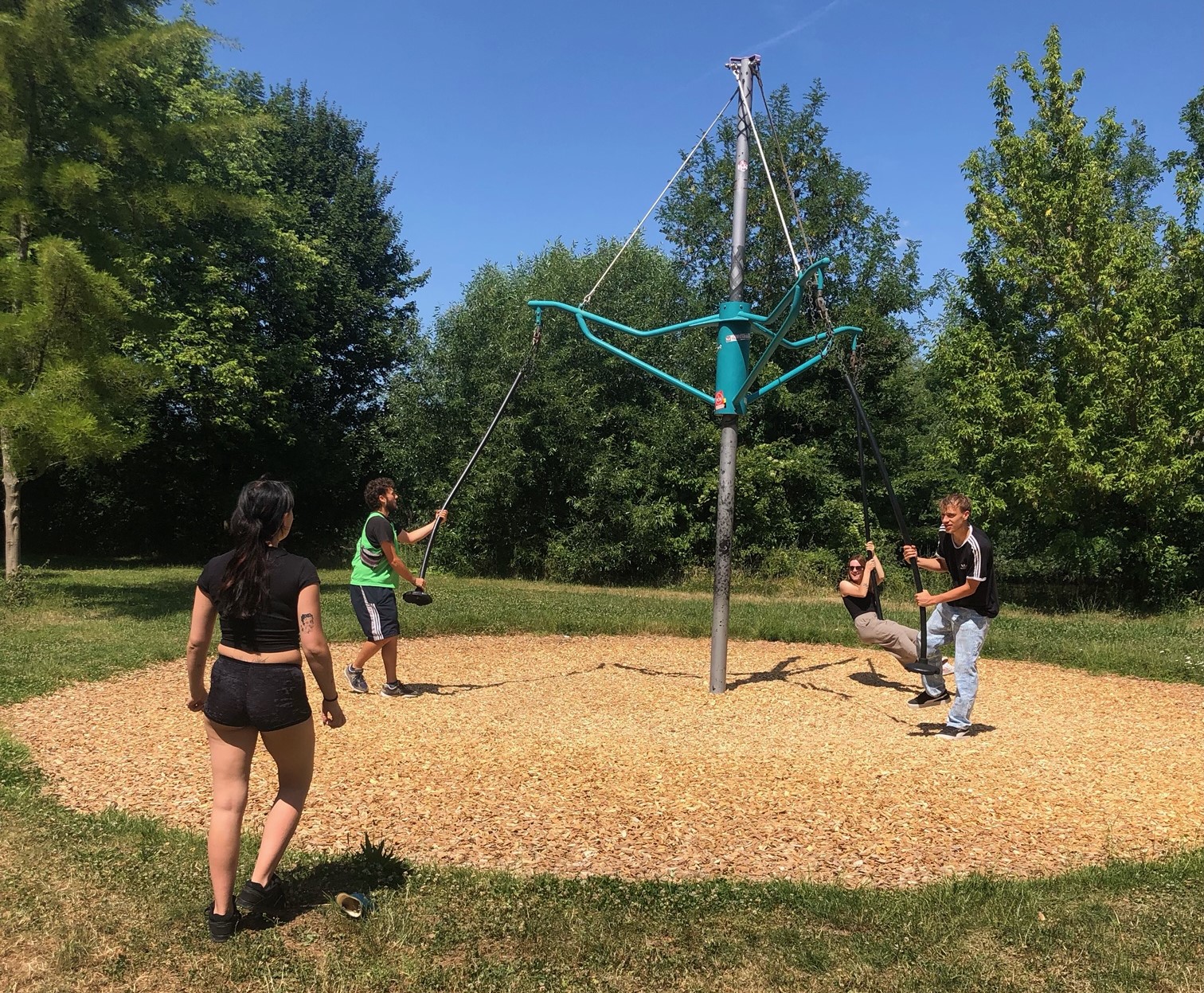 Müllsammelaktion an den Mainwiesen – Gemeinsam für eine saubere Umwelt! Drei junge Erwachsene befinden sich auf einer Schaukelkonstruktion auf einem Spielplatz, während eine Person im Vordergrund mit dem Rücken zur Kamera in Richtung der Schaukel läuft.