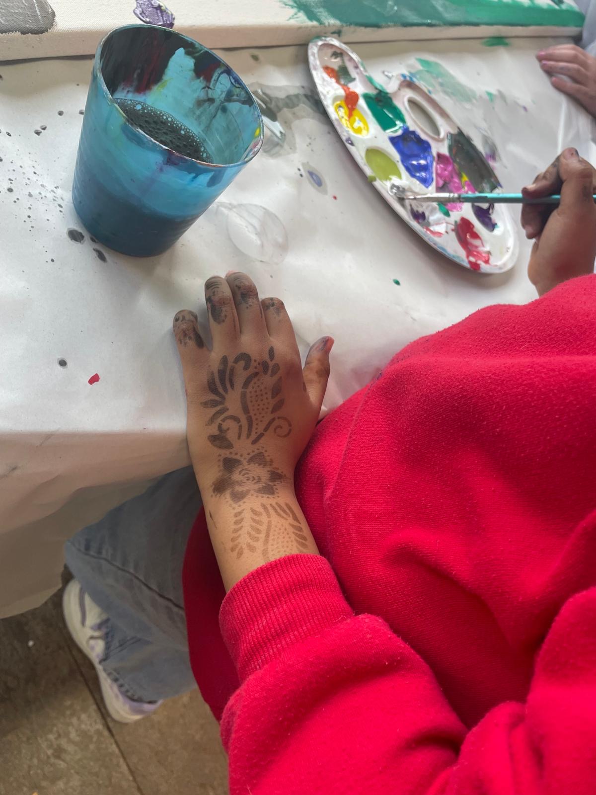 Nahaufnahme eines Kindes im roten Pullover: Die Hand ist mit Farbe verschmiert und mit einem Muster bemalt; daneben liegen ein Becher mit Wasser und eine Farbpalette.