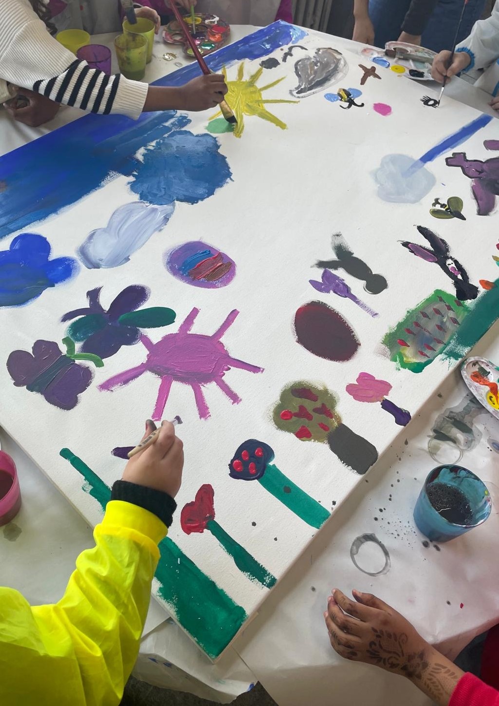 Mehrere Kinder bemalen gemeinsam eine große weiße Leinwand mit bunten Motiven wie Sonne, Wolken, Blumen und farbigen Formen.