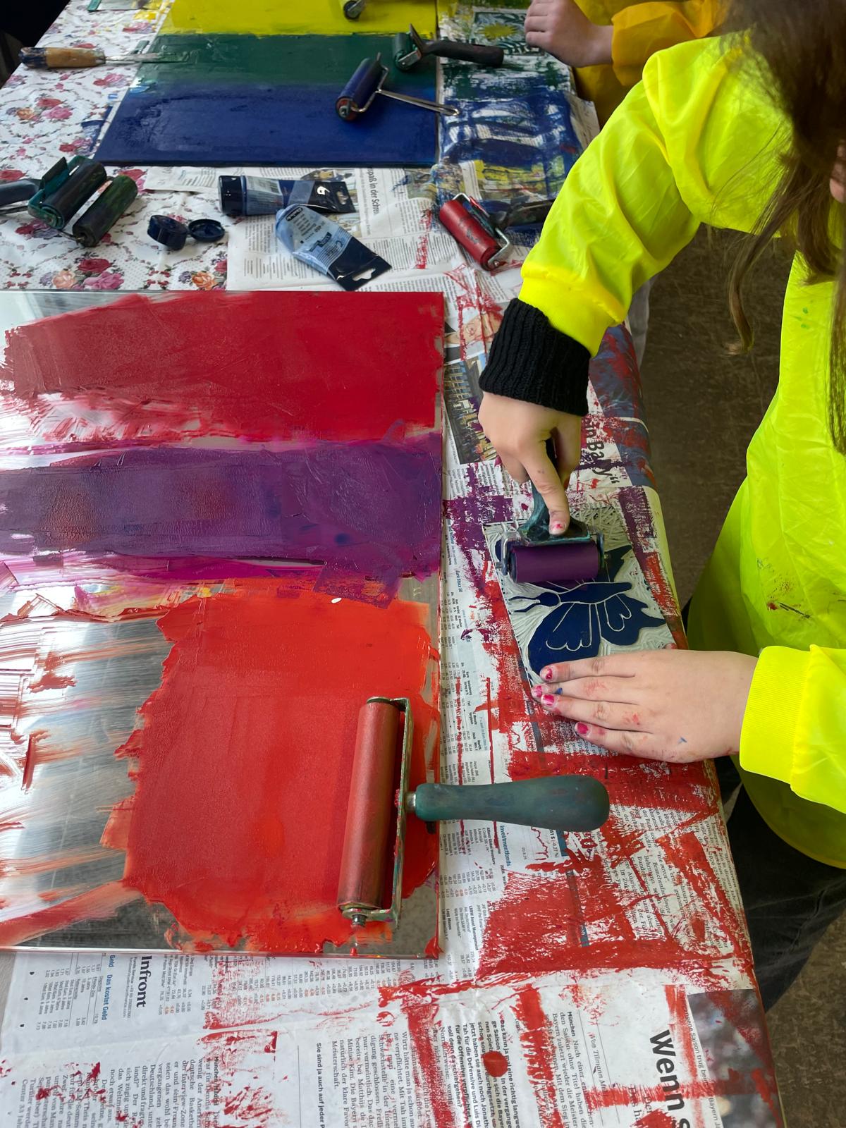 Kind in gelbem Malkittel nutzt eine Farbrolle in Lila auf einer Druckplatte; daneben liegen weitere farbige Druckplatten, Walzen und Farb­tuben auf einem mit Zeitungspapier abgedeckten Tisch.