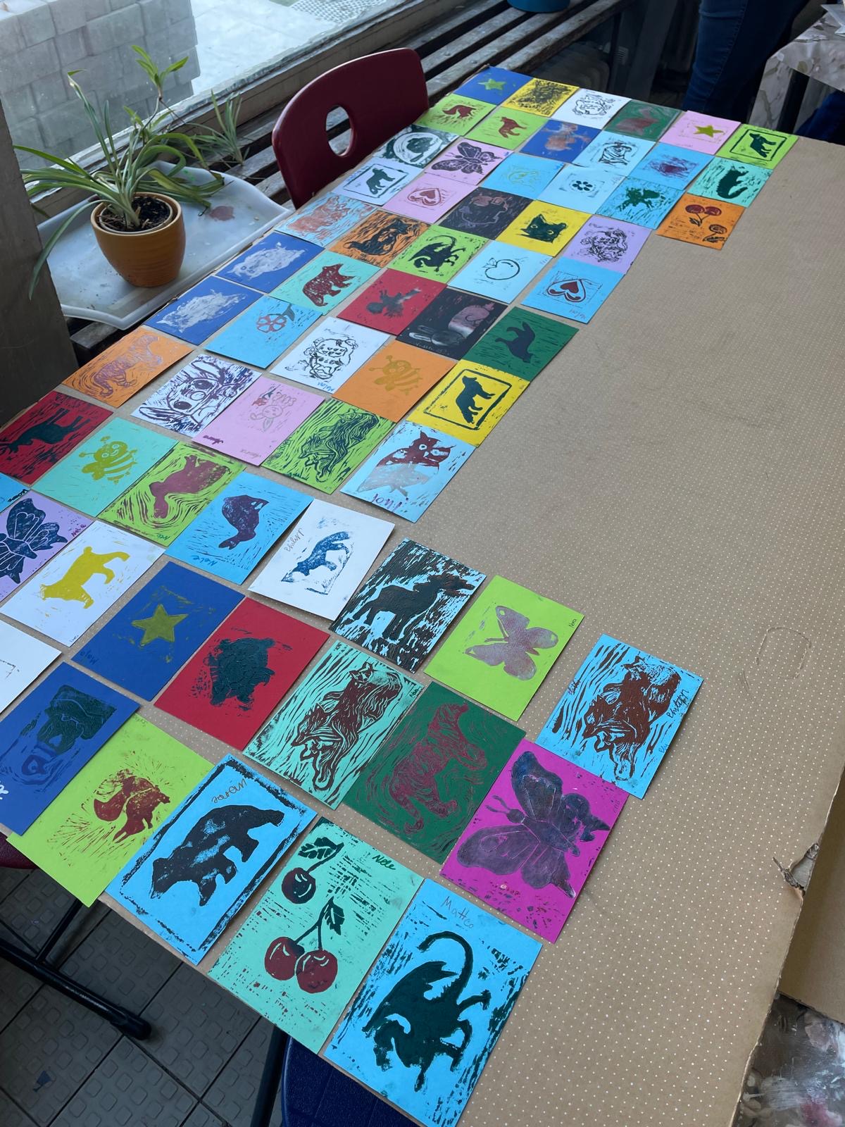 Auf einem großen Tisch sind zahlreiche bunte Linoldrucke in verschiedenen Farben ausgelegt, mit Motiven wie Tieren, Pflanzen, Sternen und Fantasiefiguren.
