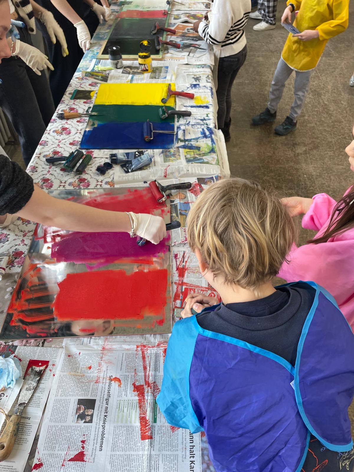 Mehrere Kinder stehen um einen langen Tisch mit Druckplatten und Farbrollen; verschiedene Farben sind aufgetragen, und es wird gemeinsam gedruckt und vorbereitet.