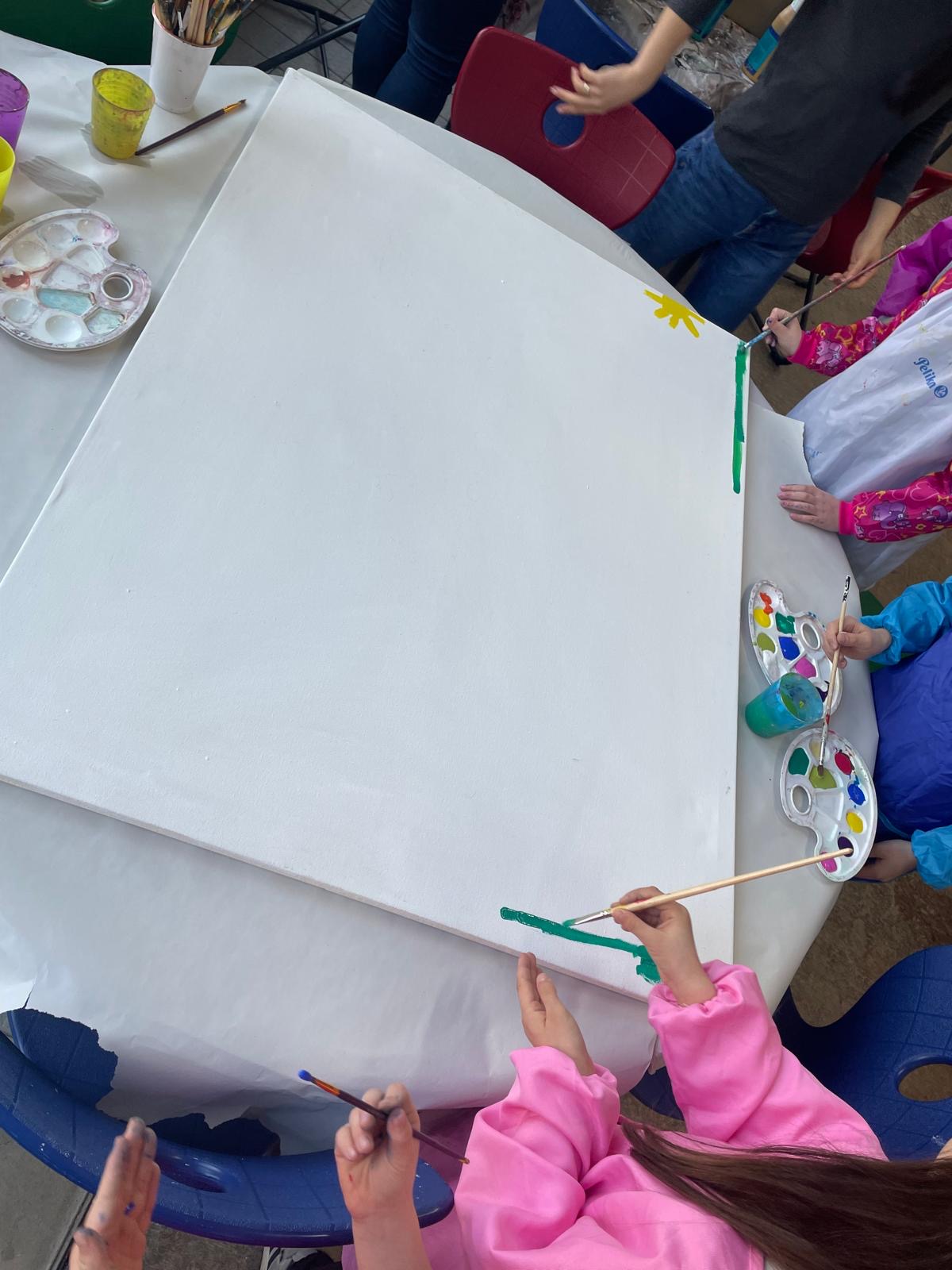 Kinder sitzen an einem Tisch und bemalen eine große, noch weitgehend leere Leinwand; am Rand entstehen erste Farbstreifen und kleine Formen, Farbpaletten liegen bereit.