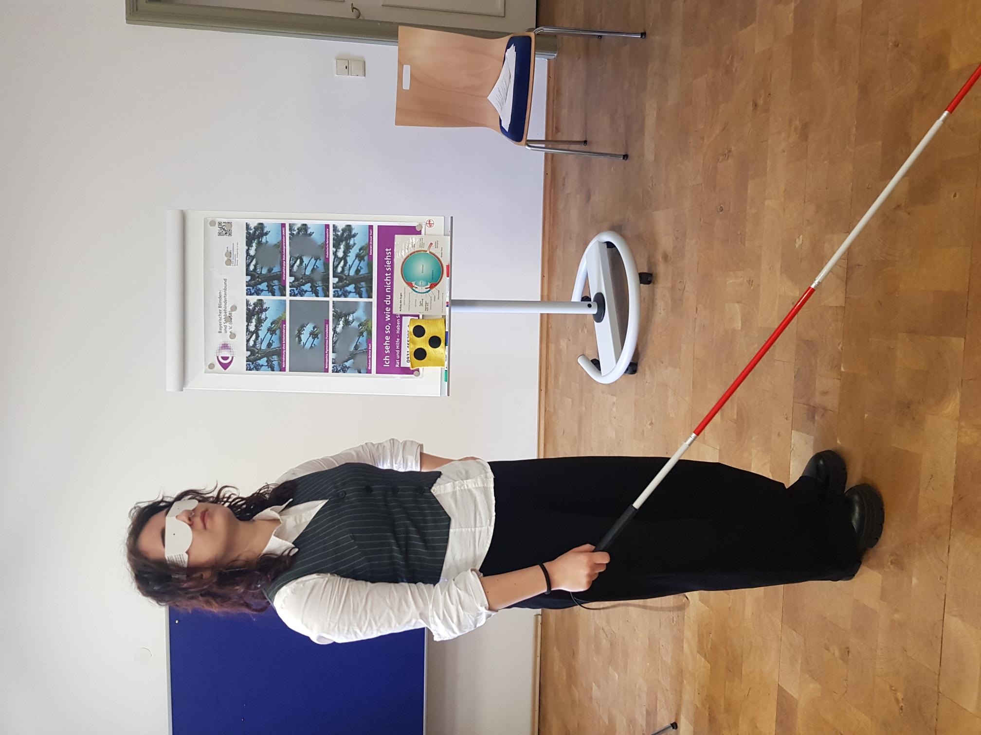 Eine junge Frau steht mit Blindenstock und Papierbrille in einem hellen Klassenraum vor einer Infotafel über Sehbehinderung.