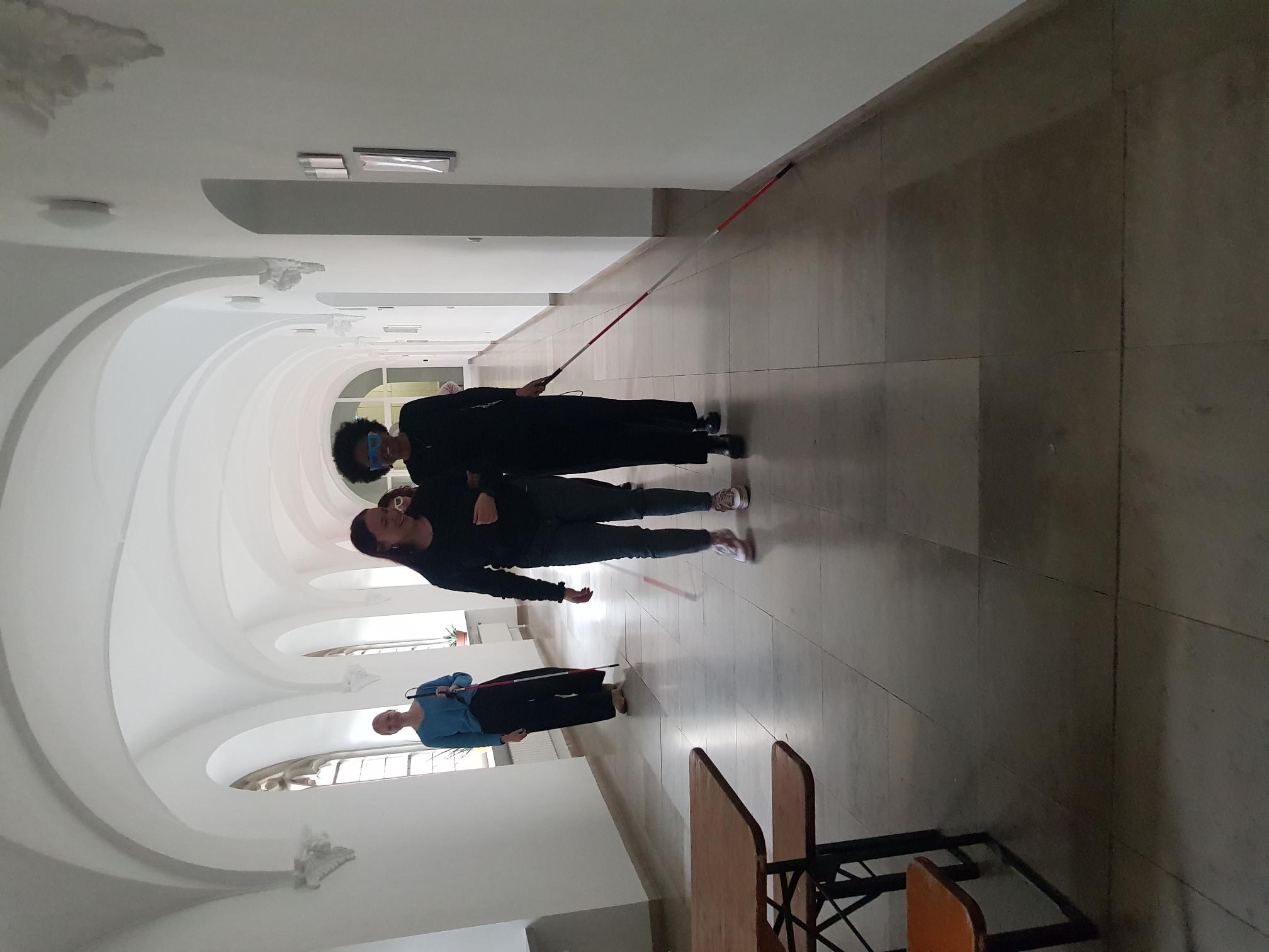 In einem hellen, gewölbten Flur mit großen Fenstern gehen zwei Personen auf den Betrachter zu, eine Person führt die andere am Arm; die geführte Person hält einen langen roten Blindenstock, während im Hintergrund weitere Menschen im Gang stehen.