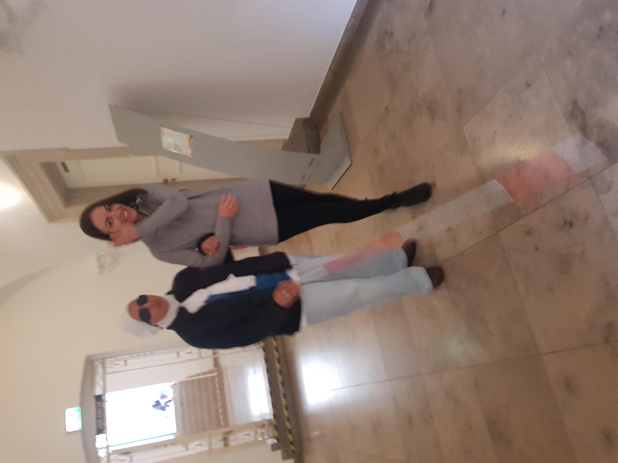 In einem hellen Flur mit Steinboden führt eine junge Frau eine andere Frau am Arm, die Blindenstock und Sonnenbrille trägt.