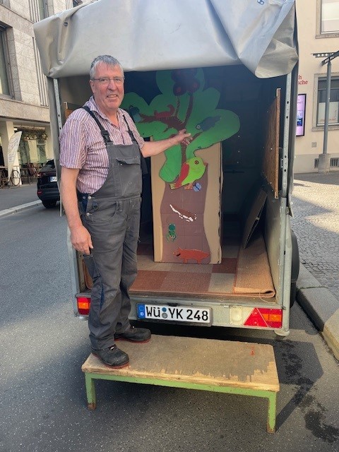 Ein Mann in Arbeitskleidung steht vor einem geöffneten Anhänger und zeigt auf einen großen, bunt gestalteten Pappbaum, der für den Transport eingeladen wurde.