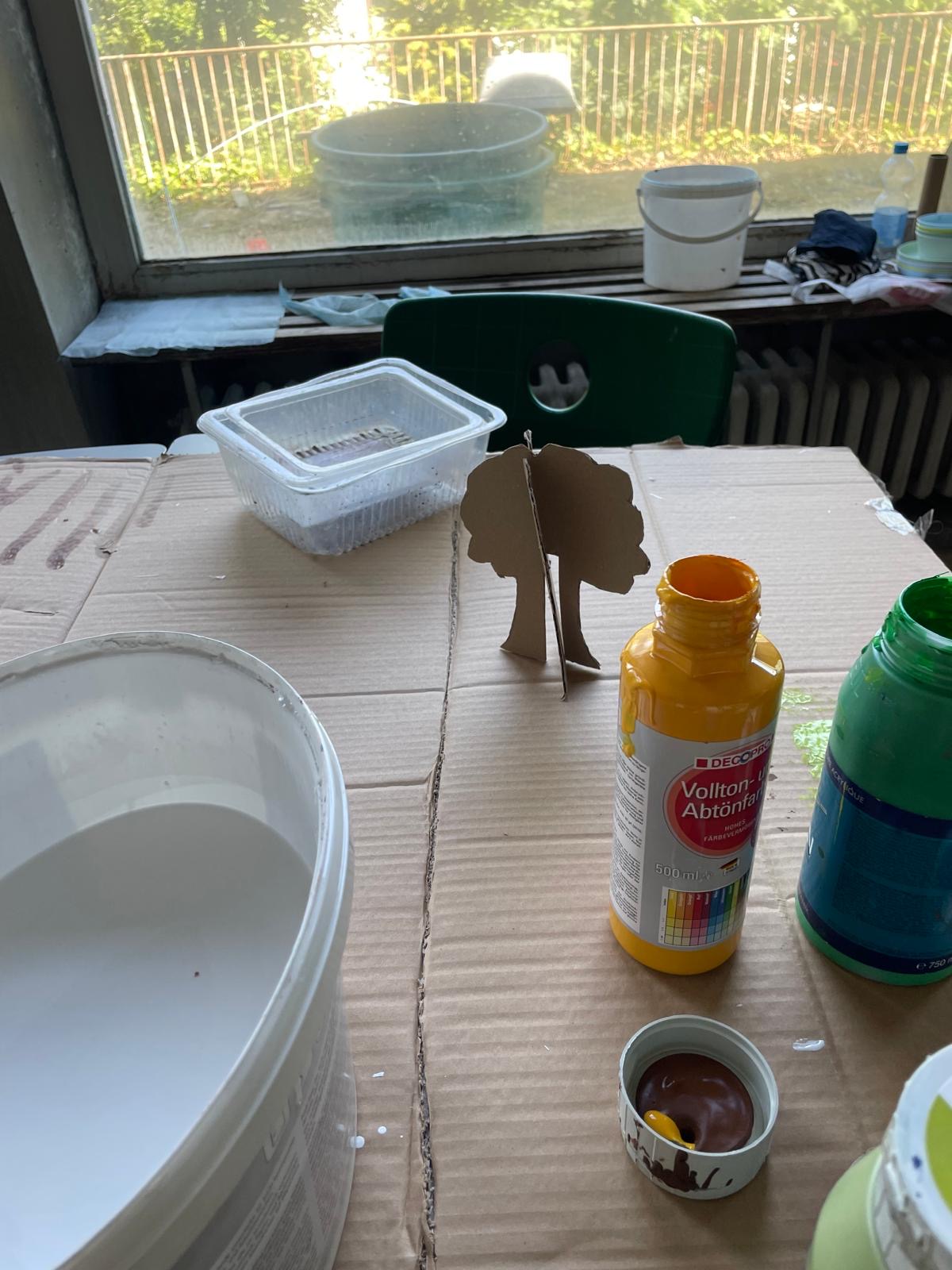 Arbeitsplatz mit einem kleinen, noch unbemalten Pappbaum sowie geöffneten Flaschen mit gelber und grüner Farbe.