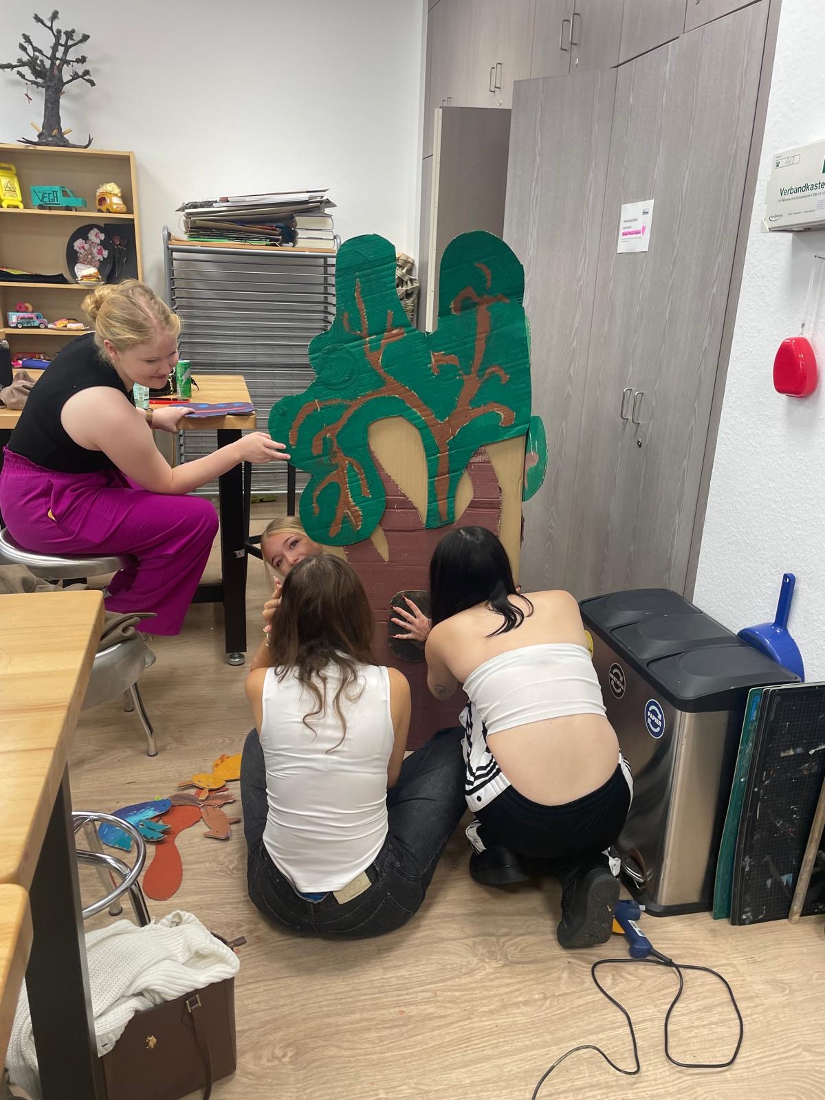Mehrere junge Frauen arbeiten gemeinsam in einem Werkraum an einem großen, grün und braun bemalten Baum aus Pappe.