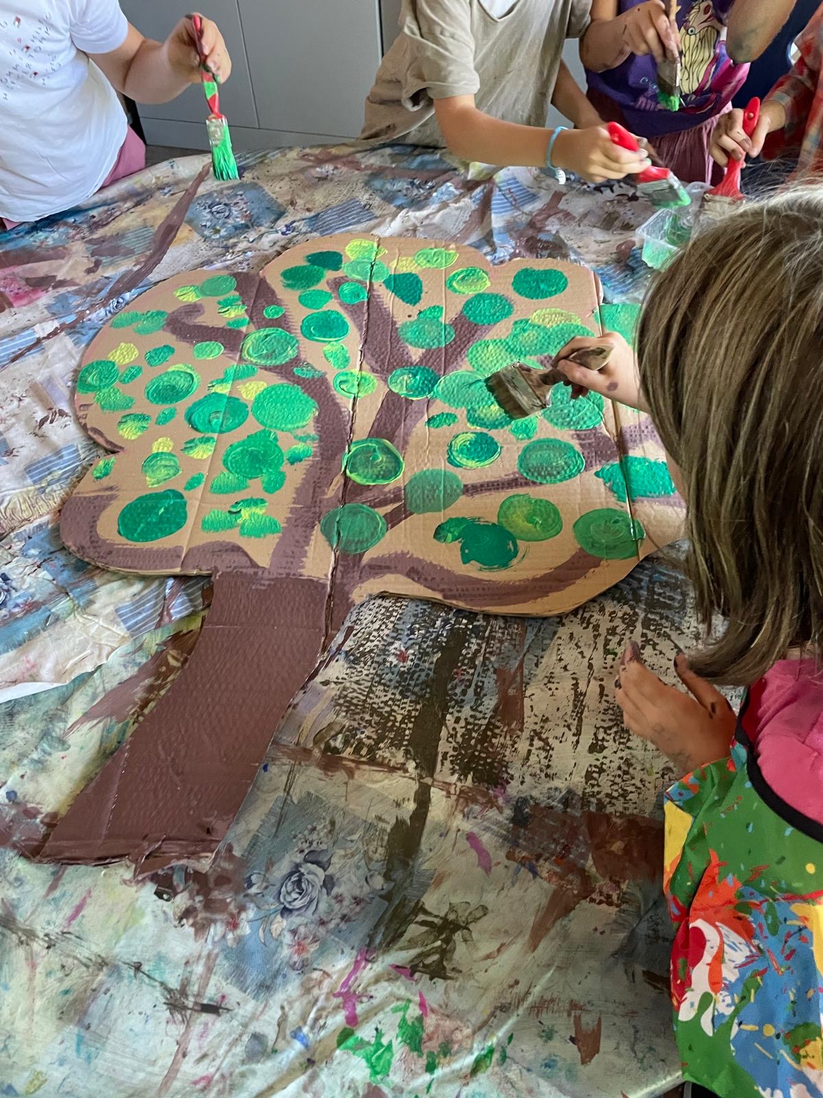 Kinder gestalten gemeinsam die Krone eines Pappbaums und malen mit Pinseln verschiedenfarbige grüne Blätter auf.