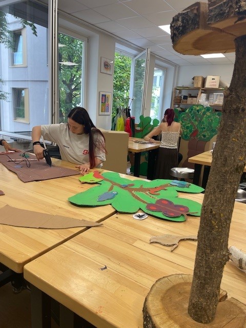 Mehrere Personen gestalten in einem Werkraum große, farbenfrohe Bäume aus Pappe; im Vordergrund liegen bemalte Baumteile auf einem Holztisch.