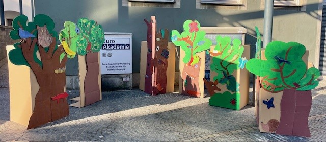 Zahlreiche bunt bemalte Bäume aus Pappe stehen vor dem Eingang der Euro Akademie Würzburg und sind mit Tieren, Pflanzen und weiteren Naturmotiven verziert.