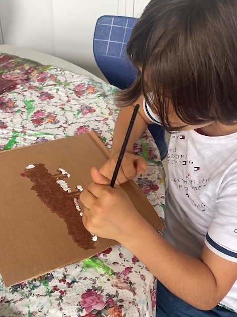 Ein Kind bemalt eine braune Pappfläche mit weißer und brauner Farbe, während es an einem mit Blumenmuster bedeckten Tisch sitzt.