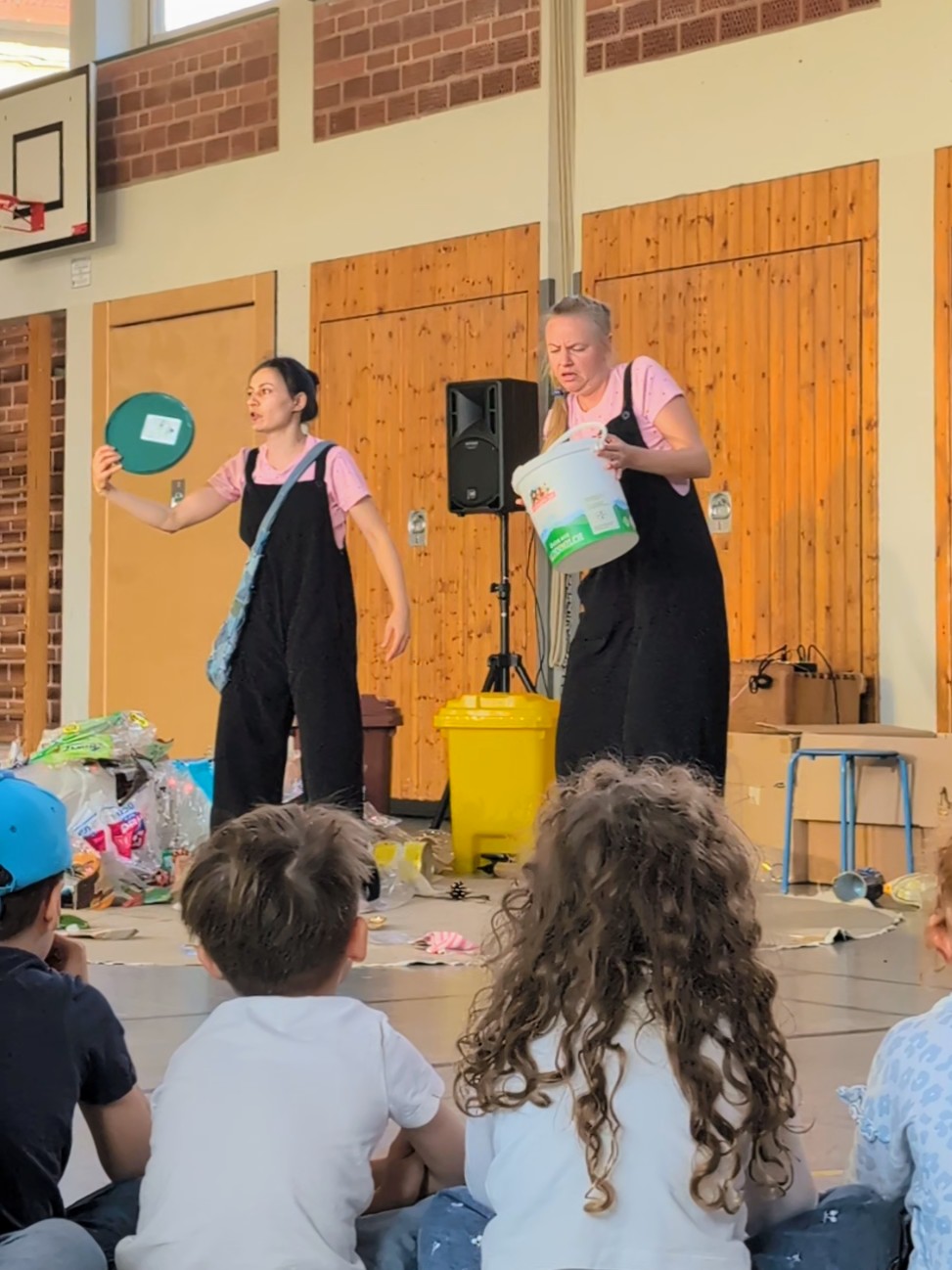 Zwei Personen führen vor sitzenden Kindern in einer Turnhalle eine Theaterszene mit Müllbehältern und Abfällen auf.