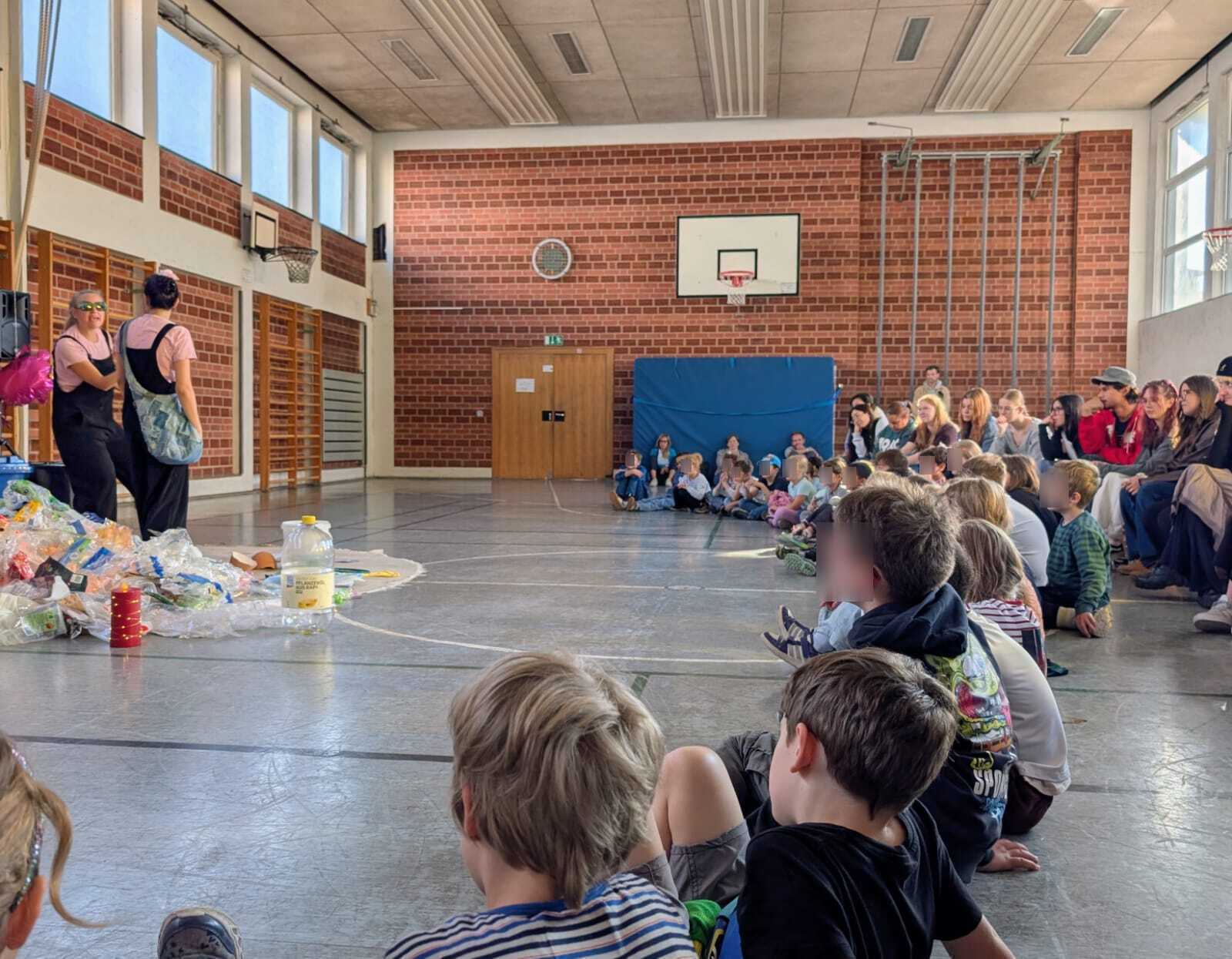 In einer Turnhalle sitzen Kinder und Erwachsene im Halbkreis. Sie schauen sich ein Theaterstück mit zwei Personen an, die neben einem Müllberg stehen.