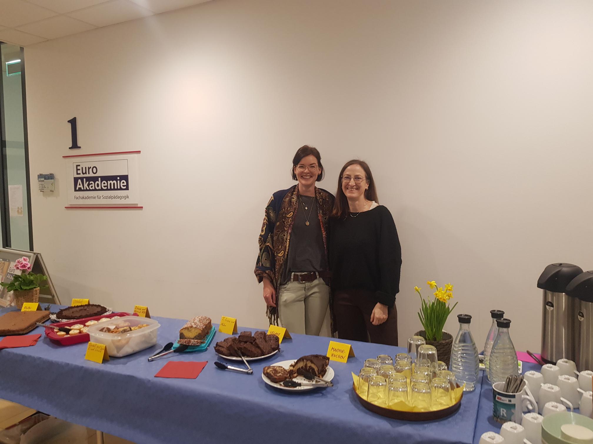 Zwei freundlich lächelnde Frauen hinter einem reich gedeckten Büffet mit verschiedenen Kuchen.