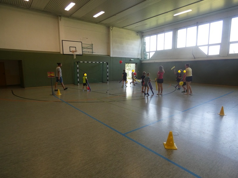 Kinder und Auszubildende spielen in der Turnhalle ein Bewegungsspiel mit Luftballons und Schaumstoffschlägern.